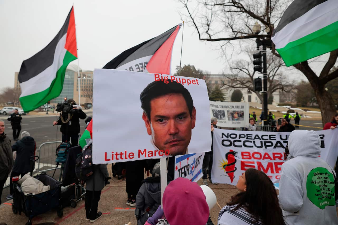 A protester holds a sign with a politician’s face labelled “Big Puppet, Little Marco” amid Palestinian flags and demonstrators gathered at a rally near a government building.