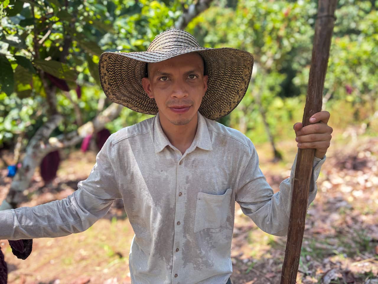A farmer in a broad-brimmed hat and linen shirt holds a large wooden stick. Behind him is a field of green trees.