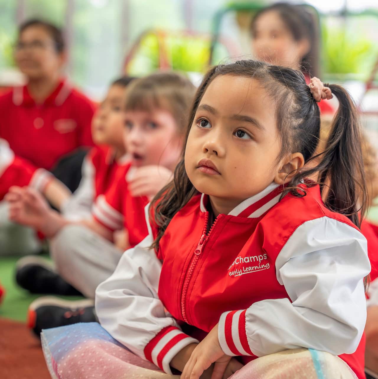 A young girl looks up at a teacher while sitting on the floor of a preschool.