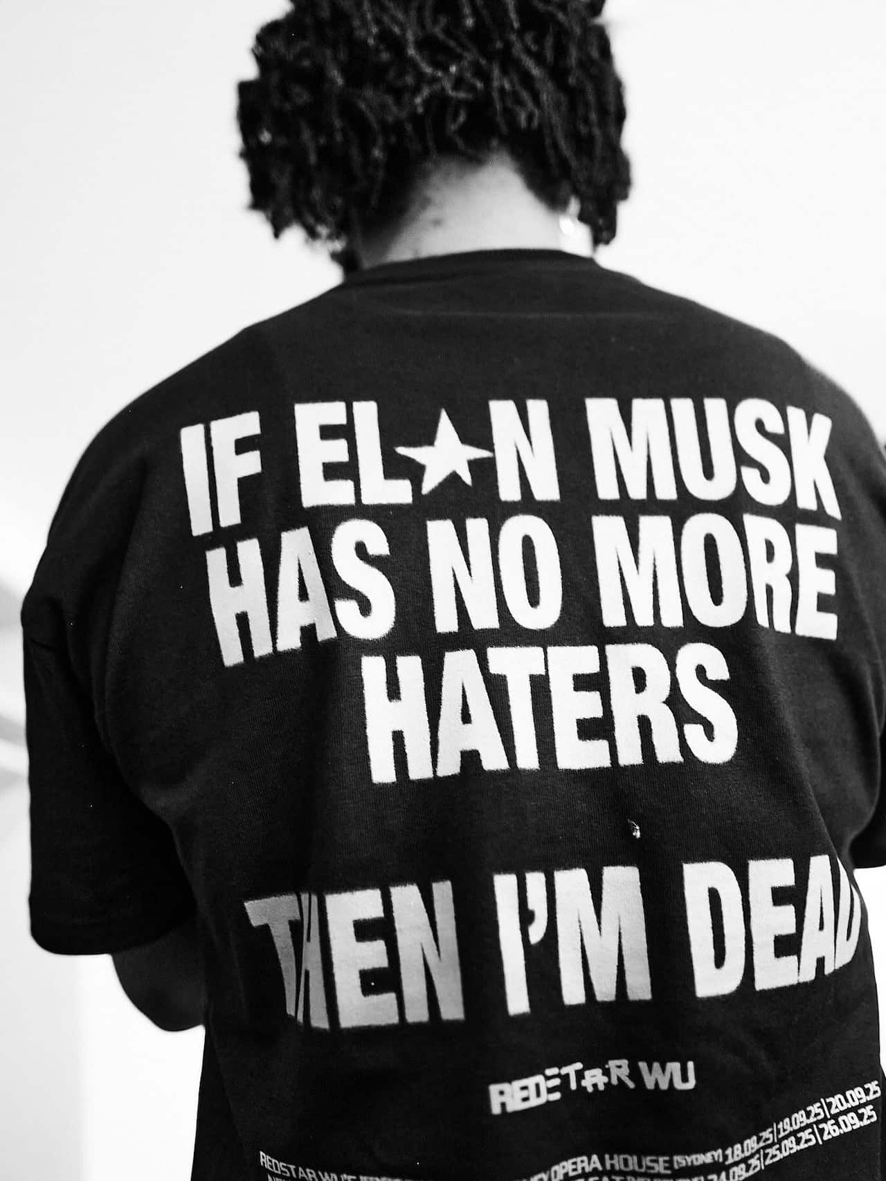 A man is facing away wearing a black t-shirt with large white writing that reads "if elon musk has no more haters, then im dead"