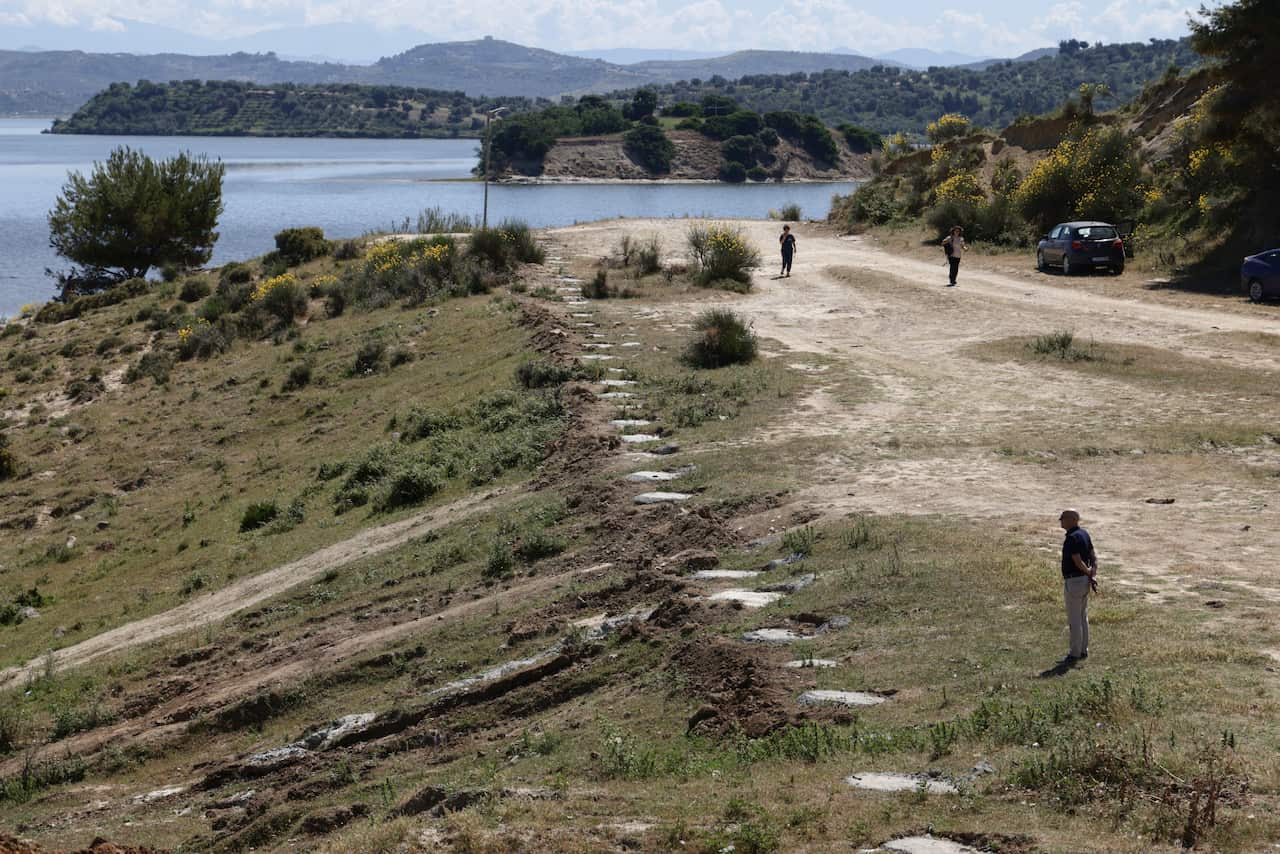 People inspect concrete foundations in a green hillside in front of a Mediterranean sea background.