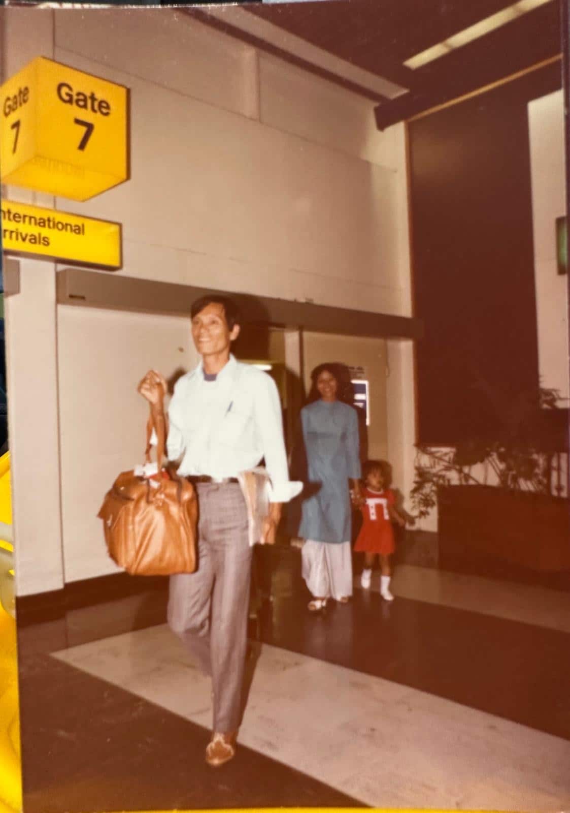 Old photograph with a man walking ahead of a woman and her daughter, at an international airport arrival door. There is a yellow-and-black sign above and in front of them that reads Gate 7, airport arrivals