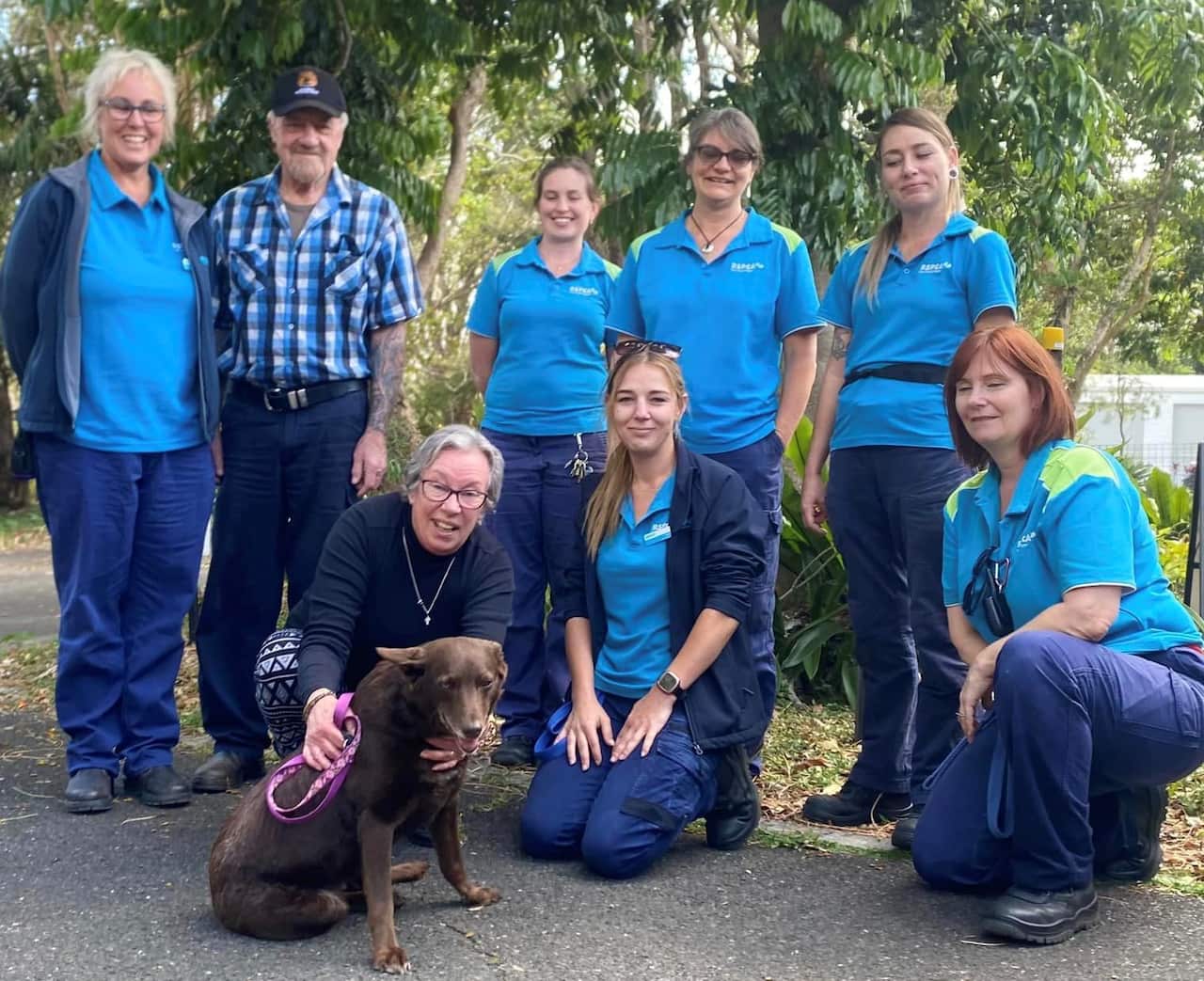 A brown kelpie dog sitting on the ground. A woman is patting the dog and there are two women kneeling next to her. Five people are standing behind.