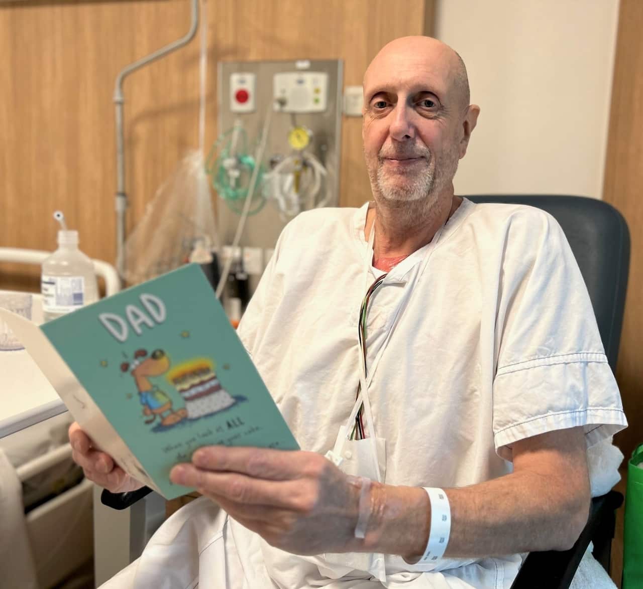 A patient in a hospital robe sits on a chair and holds up a birthday card. 