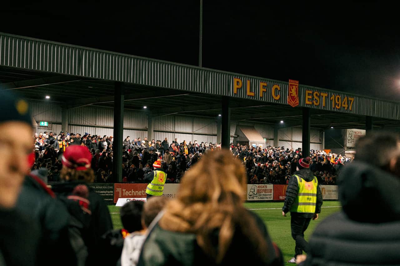 A large crowd of people stand under a soccer pavillion. 