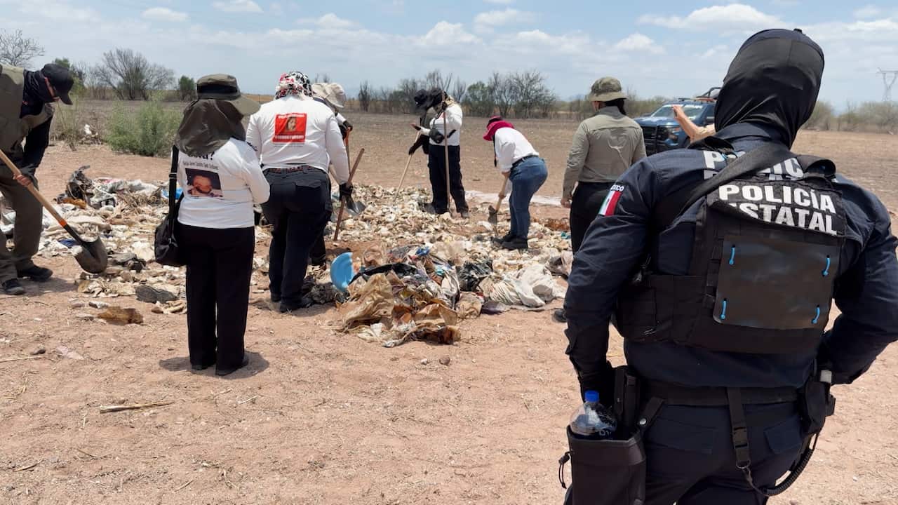 Several people wearing sun hats and long-sleeved clothing search through a pile of rubbish in a dry, open field using shovels. In the foreground, a police officer in dark protective gear observes the search.