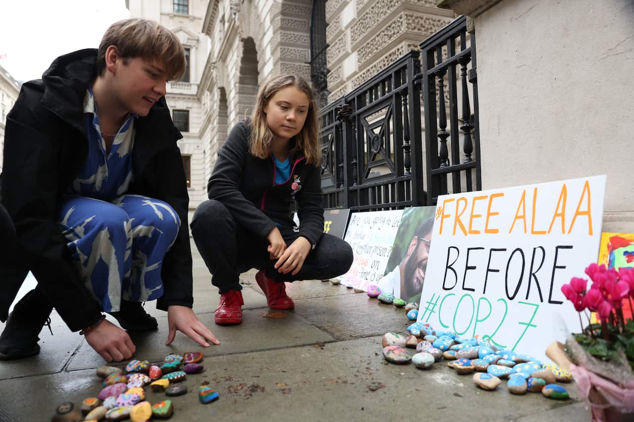 A teenage boy and girl stooping down on the pavement to look at flowers and placards placed outside a building.