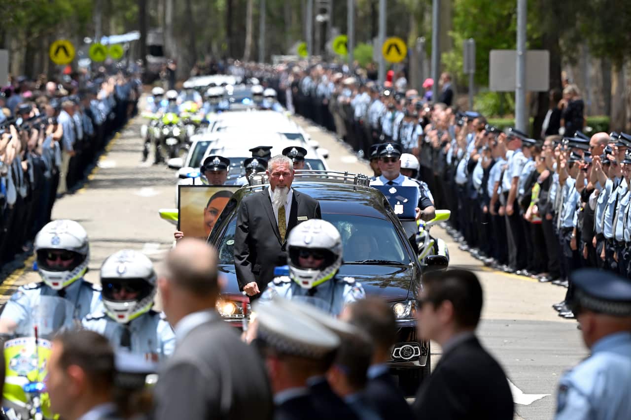 Police officers line the street where a hearse accompanied by police motorbikes drives by.