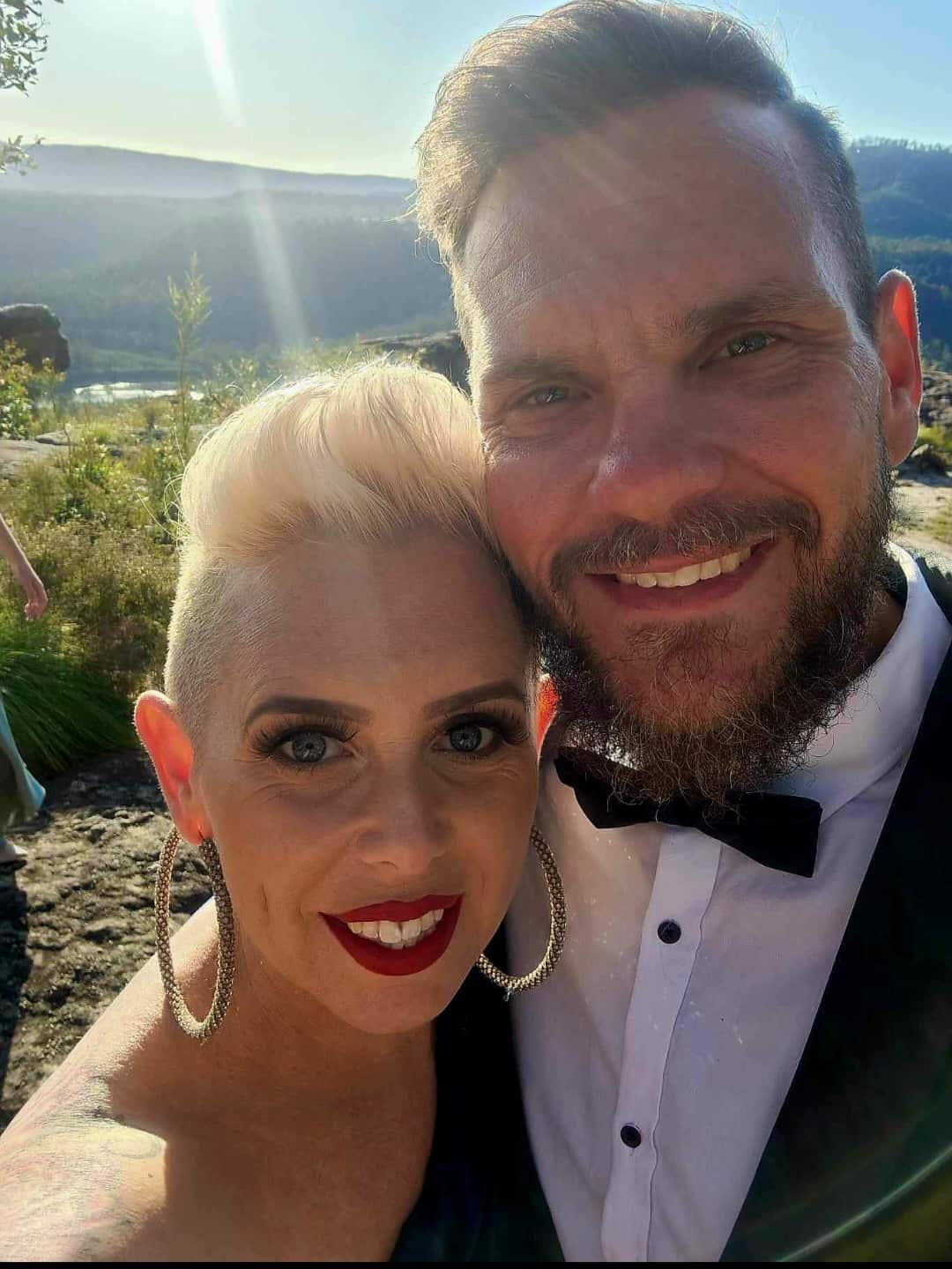 A woman wearing gold hoop earrings and a man wearing a suit pose together for a selfie in a country setting