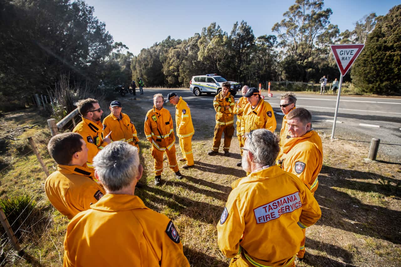 A group of firefighters standing in a circle on dry grass near a road