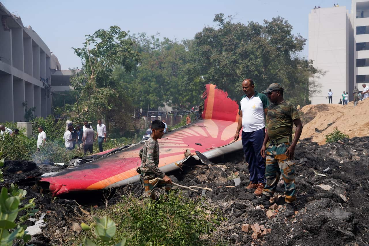 Rescue workers stand beside the wreckage of an Air India aircraft, with the broken tail section visible after the crash. Trees and a building are visible in the background.