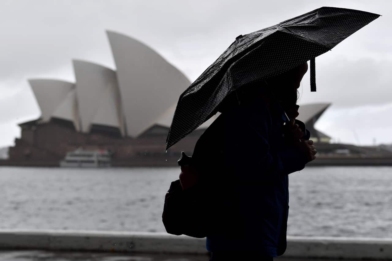 A person holding an umbrella walking. The Sydney Opera House is in the background.