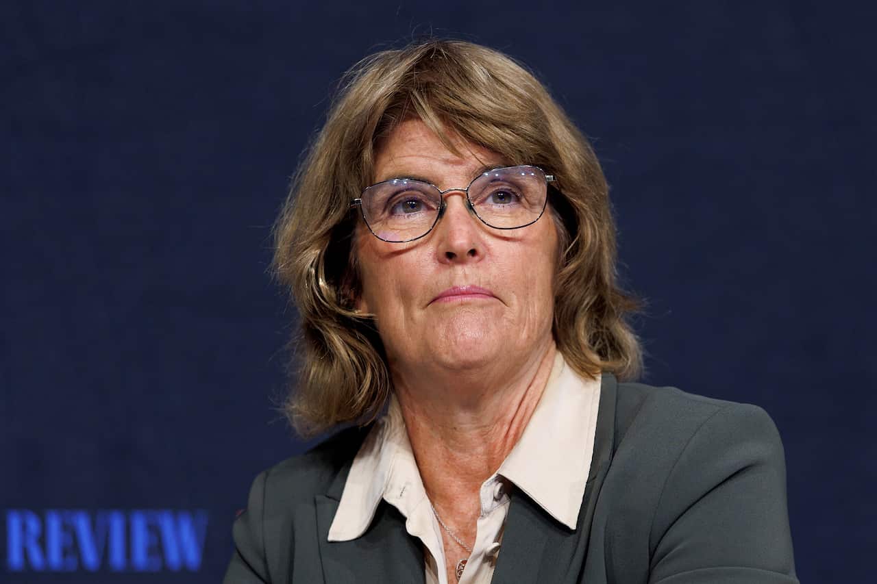 A medium shot of a woman with shoulder-length wavy brown hair and glasses, wearing a grey blazer over a light-colored collared shirt, looking upward with a thoughtful expression against a dark blue background.