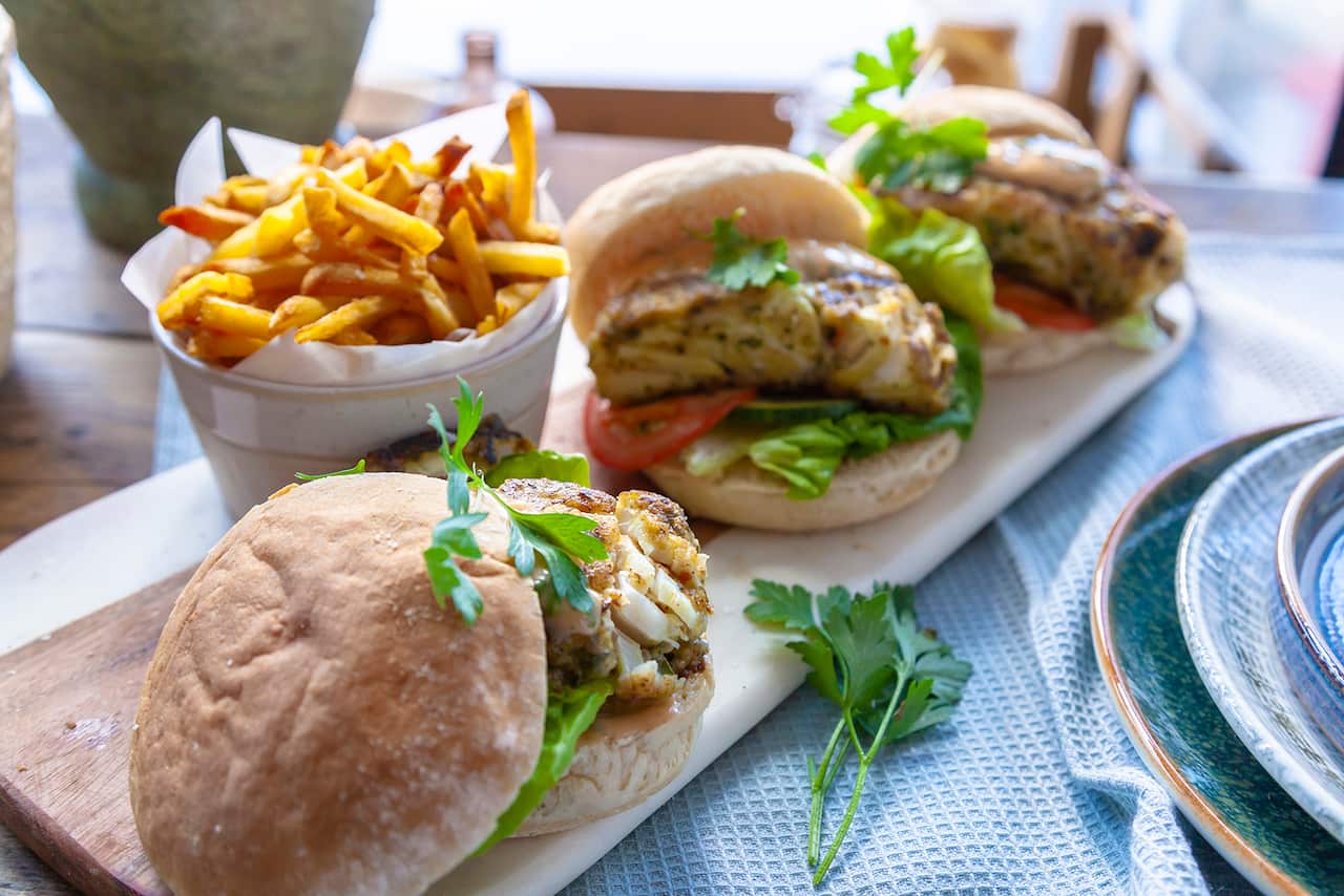 Three fish burgers in soft buns sit on a long wooden board. A small bucket with golden fries sits beside the board. 