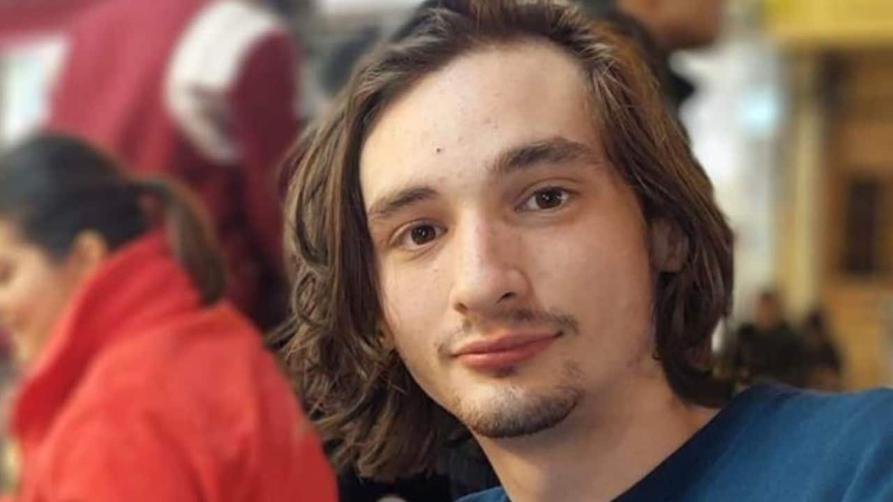 a young man with long brown hair and facial hair smiles at the camera with a woman in a red jacket blurry in the background