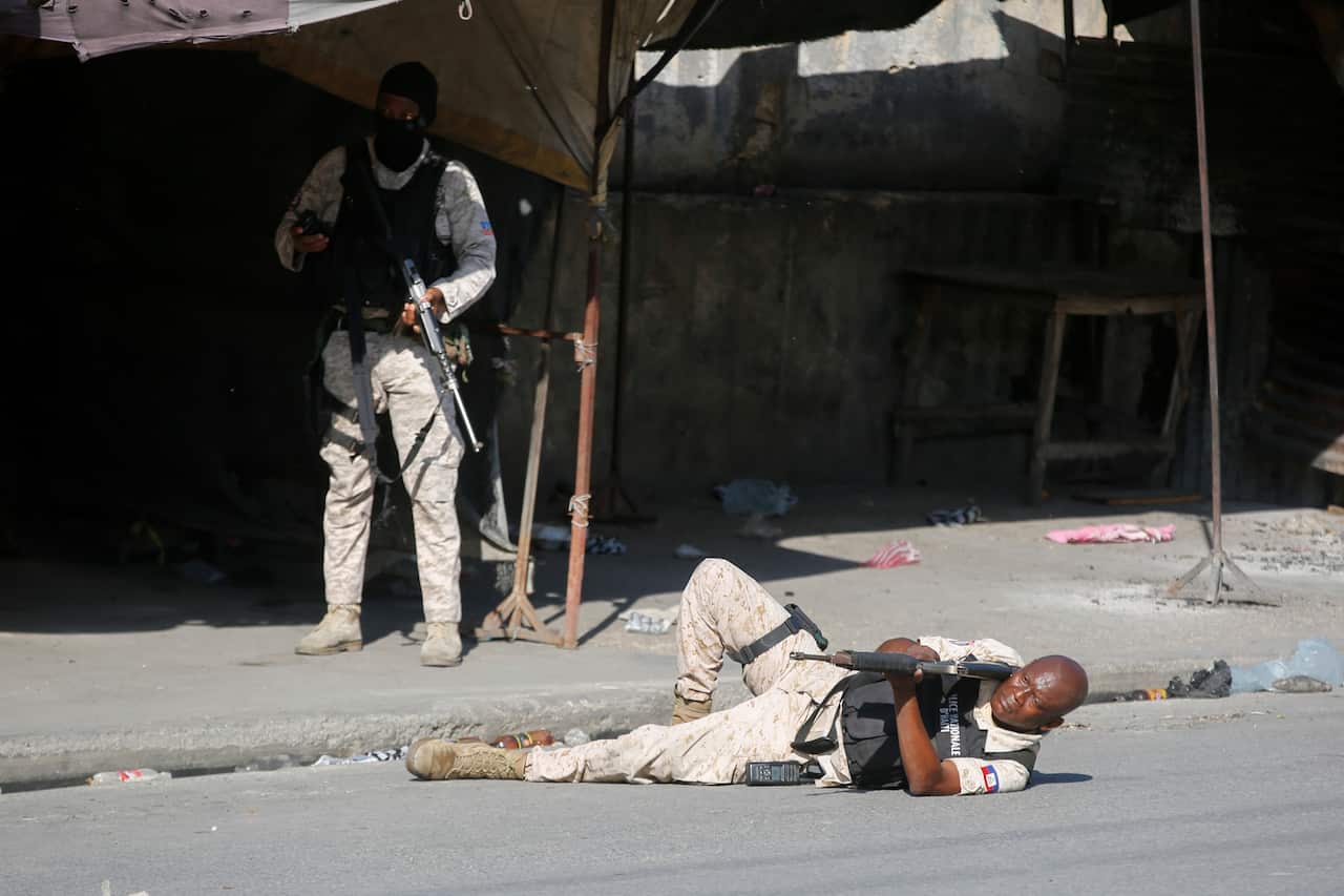 A police officer aims lies on a road aiming a rifle in the air. Another officer stands beside him.