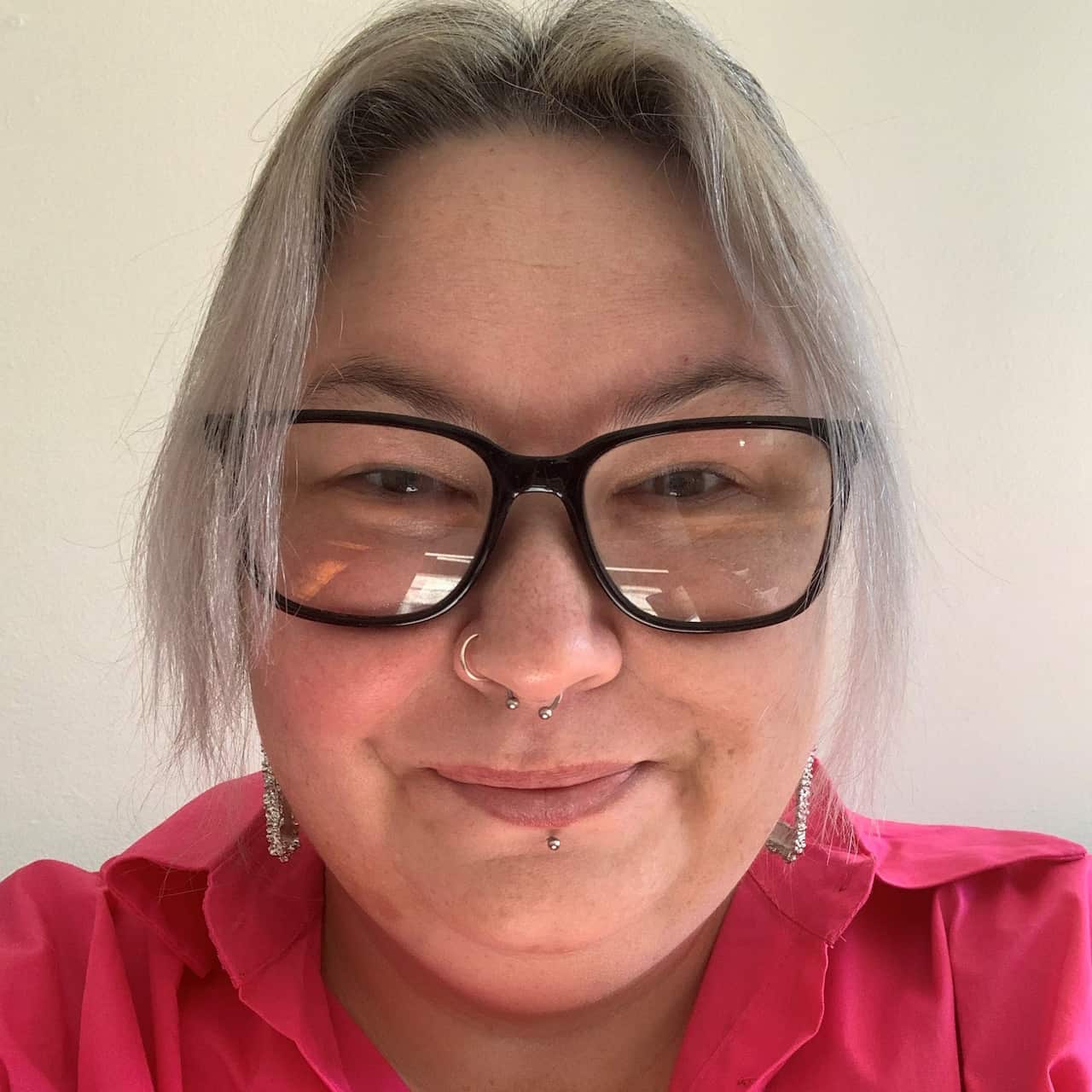 A woman with black framed glasses and facial piercings in a bright pink shirt