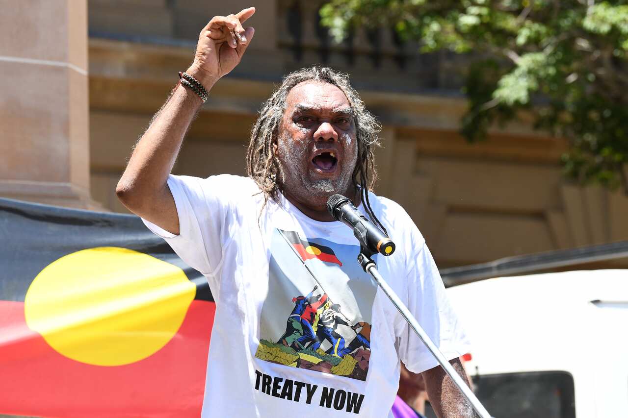 A man speaks outside from behind a microhpne. He is wearing a t shirt reading treaty now, there is an Aboriginal flag behind him