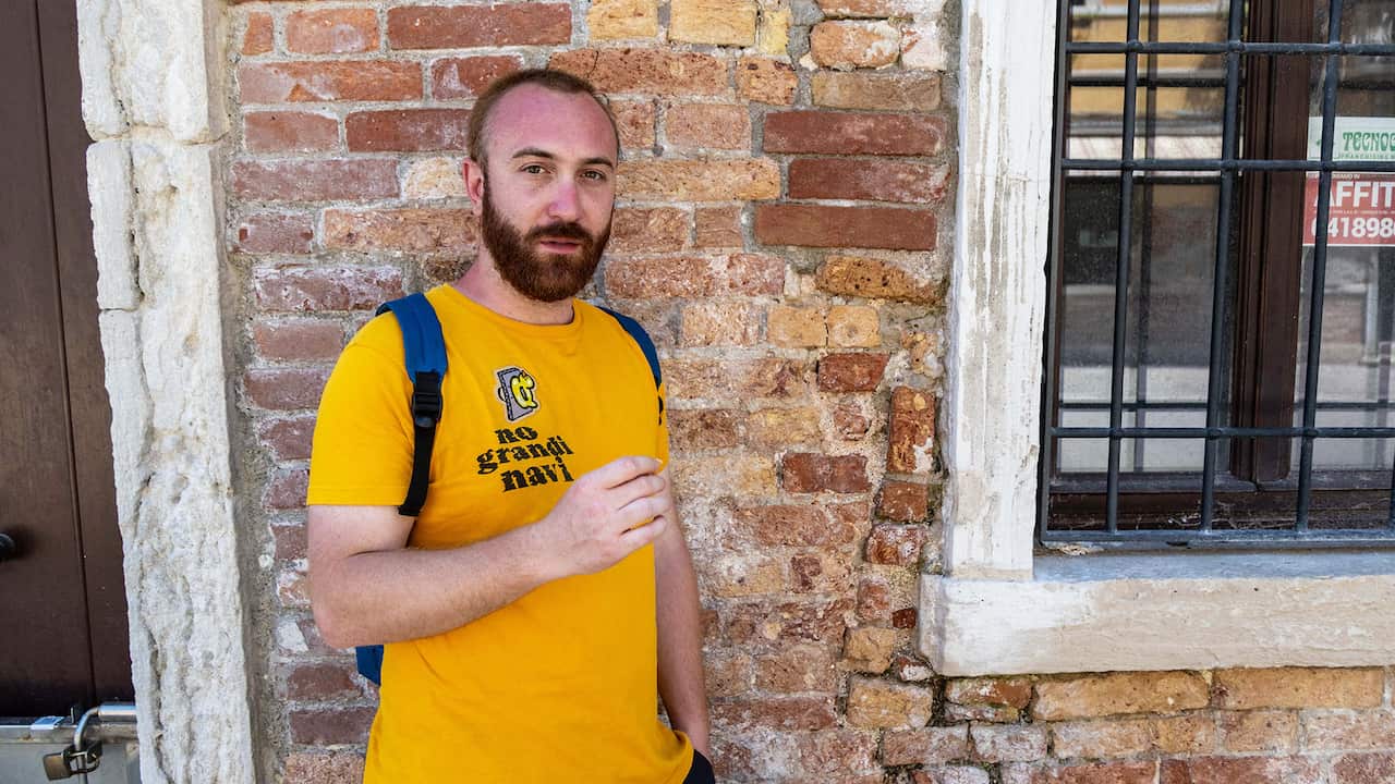 A bearded man wearing a yellow t-shirt and a backpack standing against a brick wall 