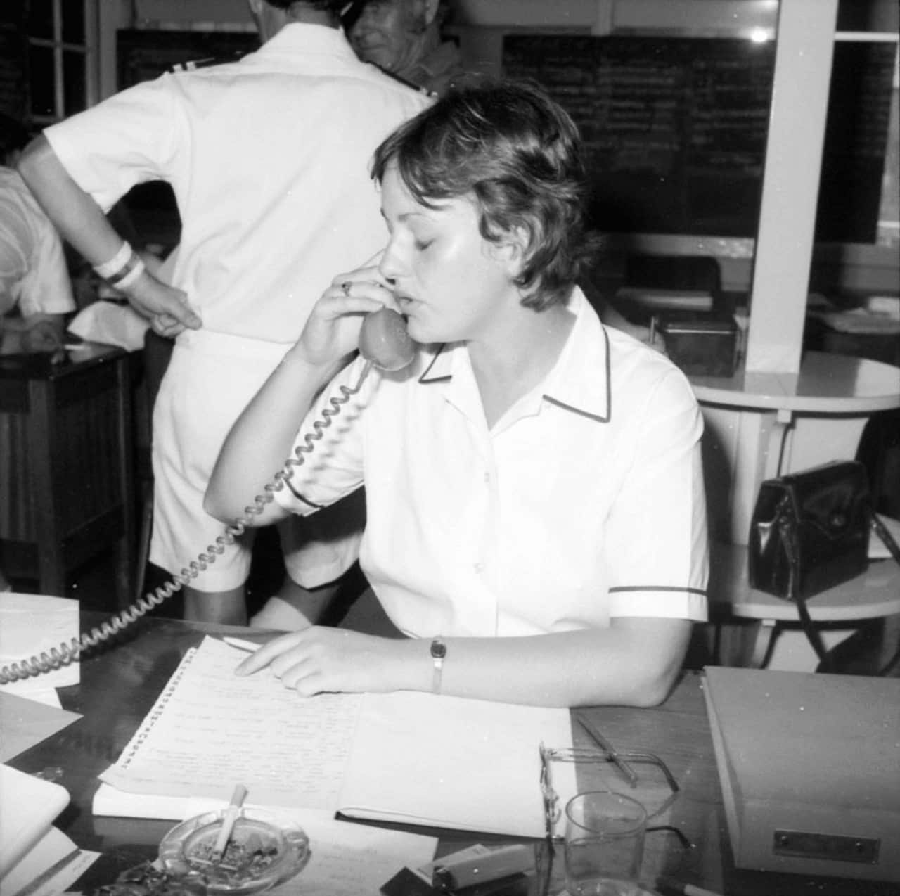 Black and white pictre of a woman in a white naval uniform answers a phone, with the chord dangling off the screen. She sits in front of a notepad, with a cigarette tray above it.