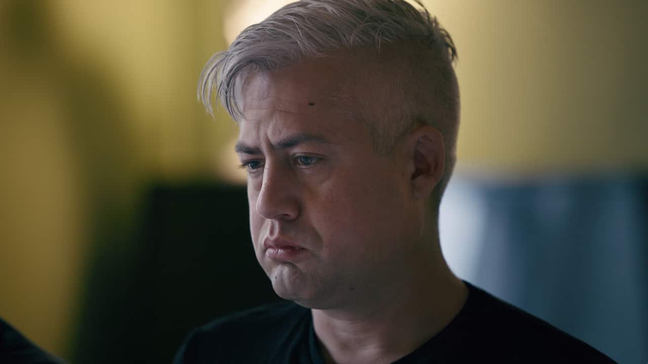 A medium close-up shows a man with short, silver-toned hair looking downward with a somber, pensive expression against a softly blurred yellow and blue background.