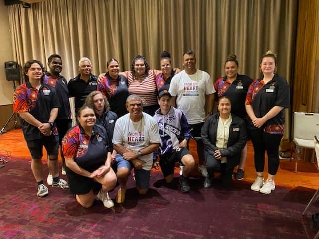 A group of young people wearing black uniforms standing in a semi circle, with two older men wearing white t shirts amongst them. 