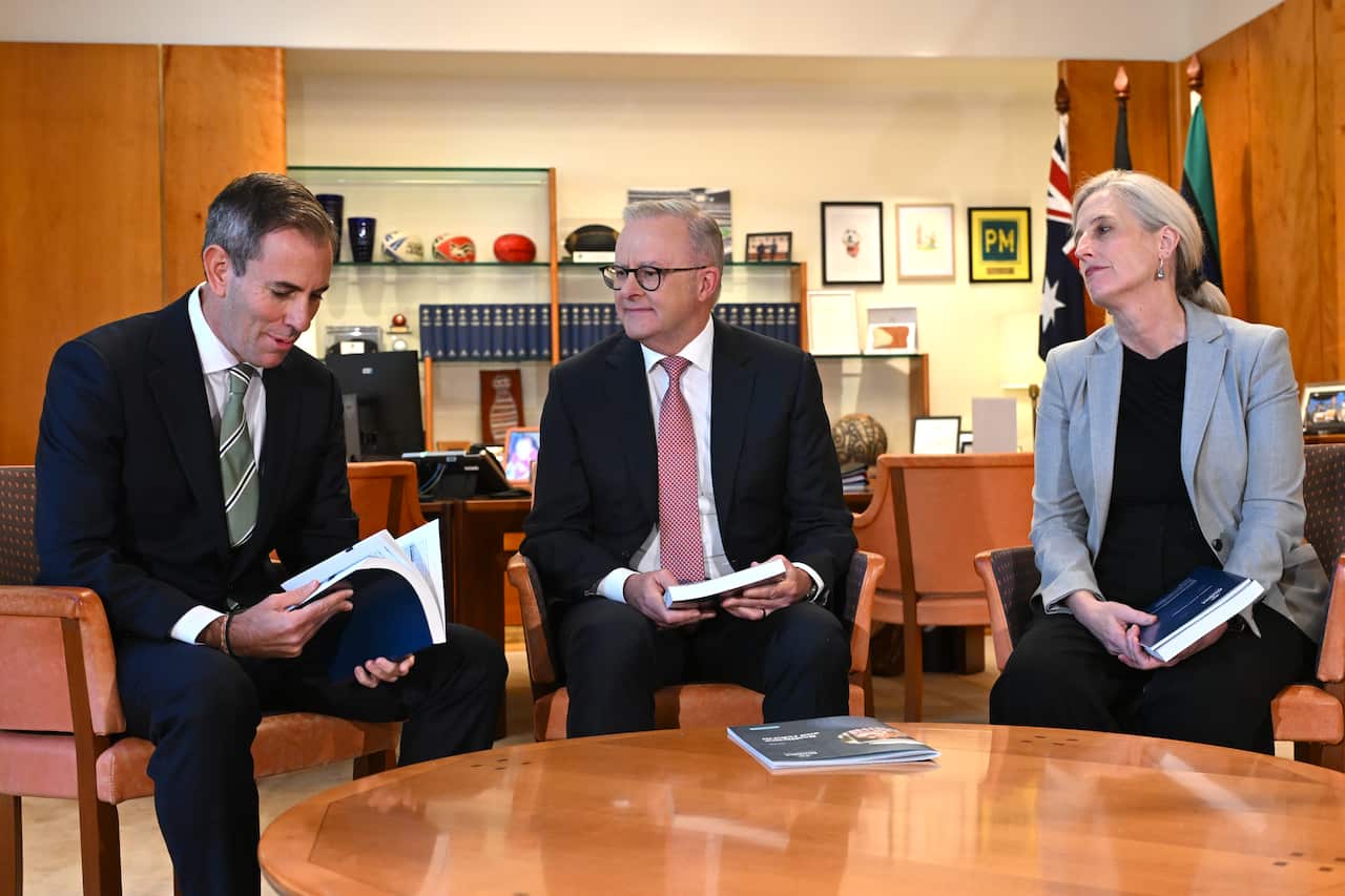 (Left to right): Jim Chalmers. Anthony Albanese and Katy Gallagher hold budget documents as they sit at a wooden round table and talk.