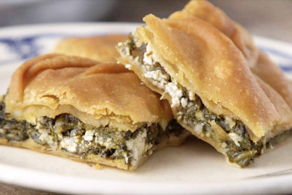 Two pieces of spanakopita, with thick golden pastry and a cheese and greens filling, sit on a blue and white plate. 