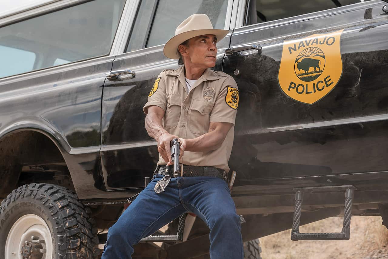 A Native American policeman crouches beside a police vehicle, gun in hand.