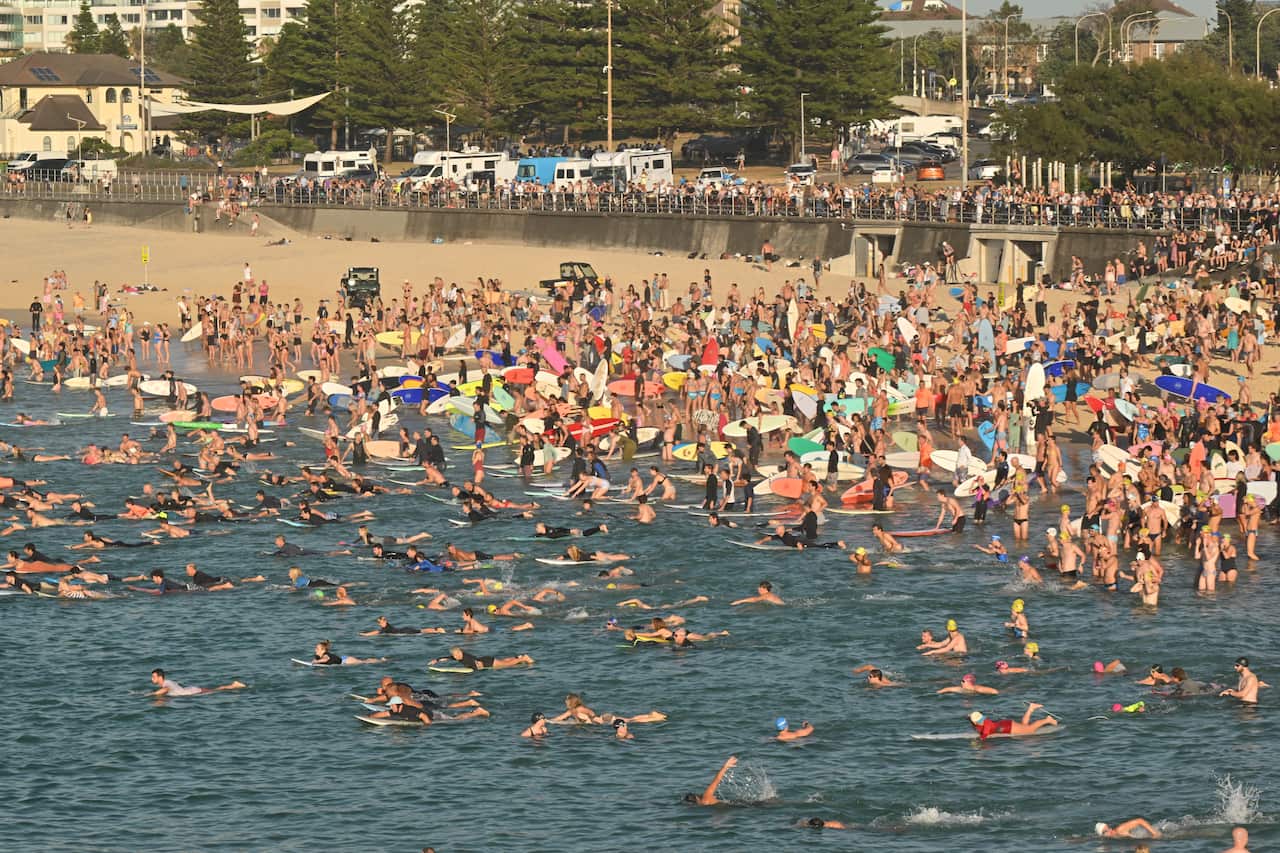 Large crowds of people on Bondi Beach, with many entering the water on boards.