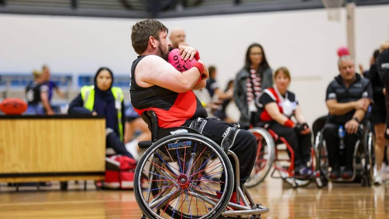 a man in a sports uniform hugging an Aussie rules ball using a wheelchair on an indoor court. There are other players and umpires blurry in the background