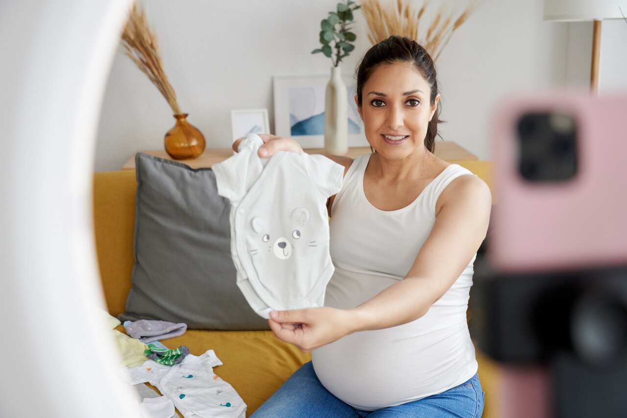 Pregnant female blogger showing baby clothes while broadcasting with mobile phone.