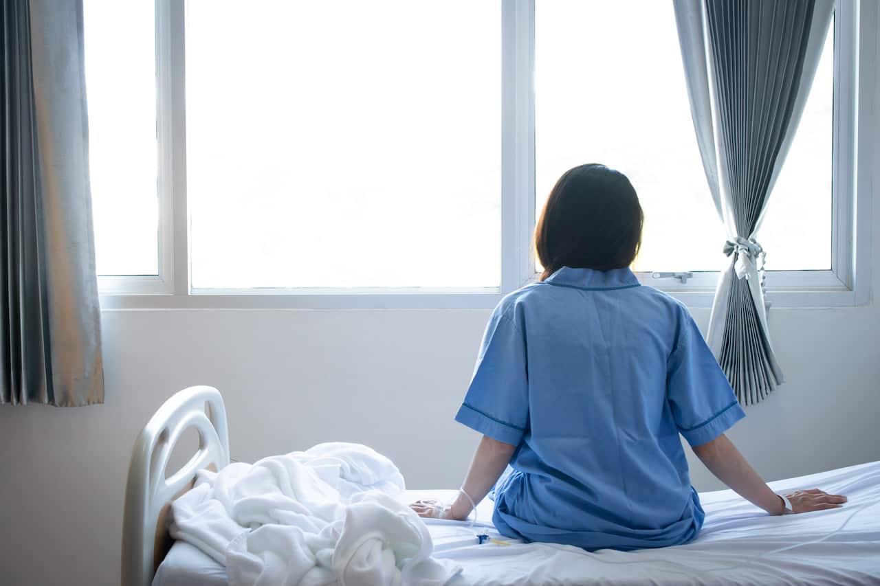 Back view of patient woman sitting on bed in hospital ward, looking away at window 