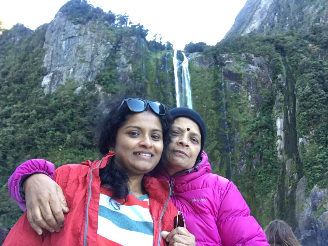 An Indian woman is embraced by her mother as the pair stand outside in jackets in front of a waterfall.