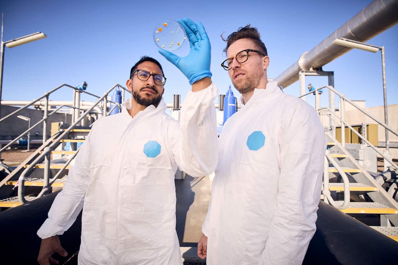 Two men in lab coats stand outdoors with metal staircases seen behind them. One holds a petri dish in a blue-gloved hand. 