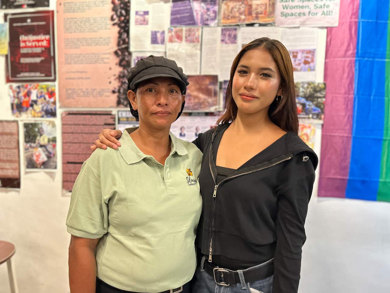 A Filipino woman in a black hat and green polo shirt has her arms around a younger woman wearing a black hoodie and jeans. They are looking into the camera with neutral facial expressions. Behind them is a wall of posters.