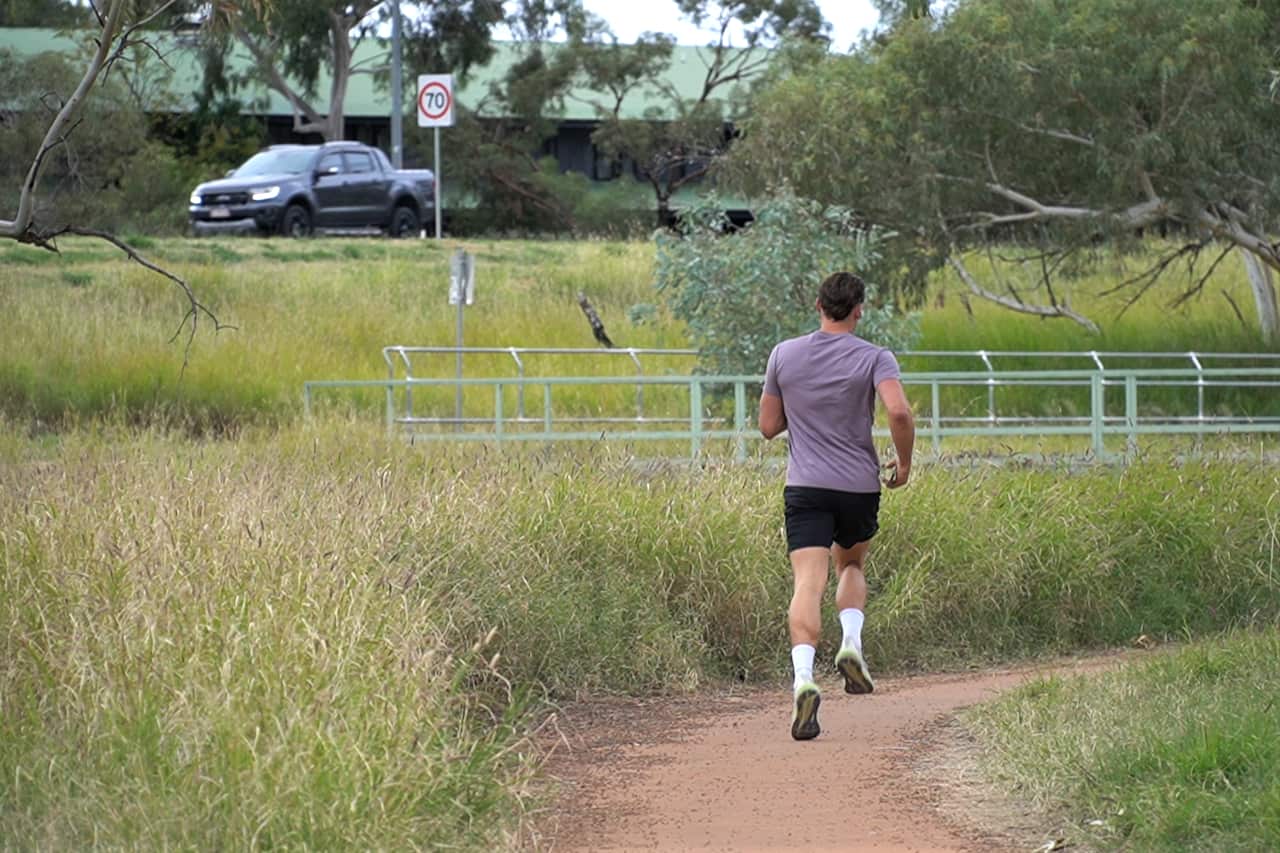 A man is running on a narrow muddy track surrounded by tall weeds.