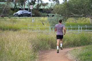 A man is running on a narrow muddy track surrounded by tall weeds.