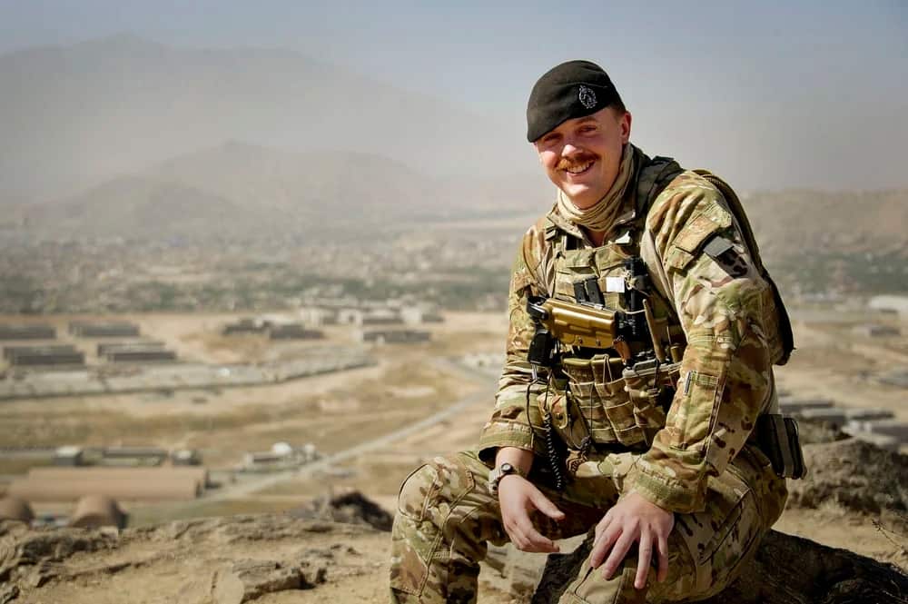 A young white man wearing an army uniform and beret sitting on the ground and smiling. A city in an arid landscape is behind him. 