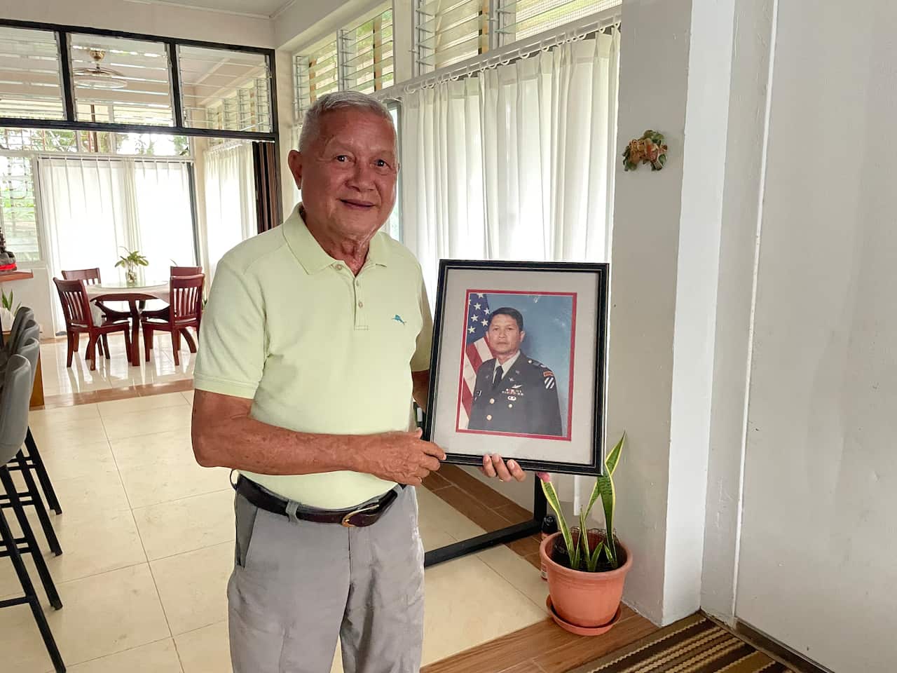 An older man wearing a polo shirt stands inside a house holding a framed portrait of a man in military uniform with an American flag in the background.