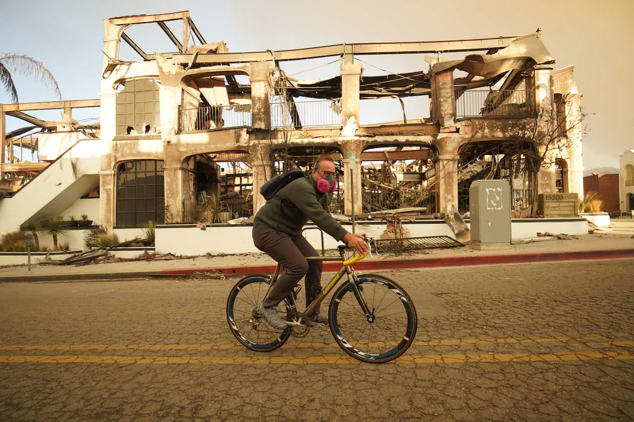 A man rides his bike past a burnt house.
