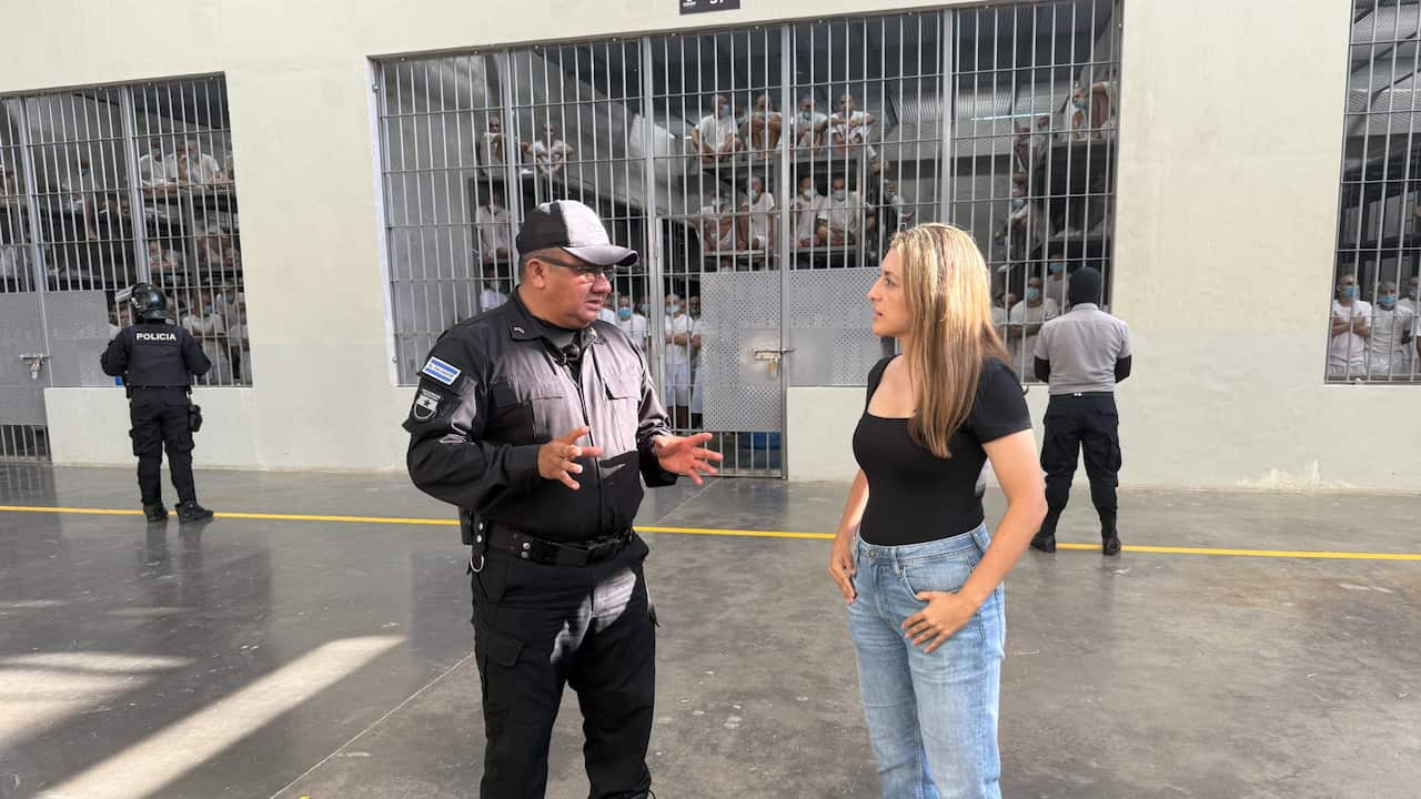 A woman wearing a black t-shirt and blue jeans is talking to a man wearing the uniform of a prison official. Security guards and cells with inmates inside are visible in the background. 