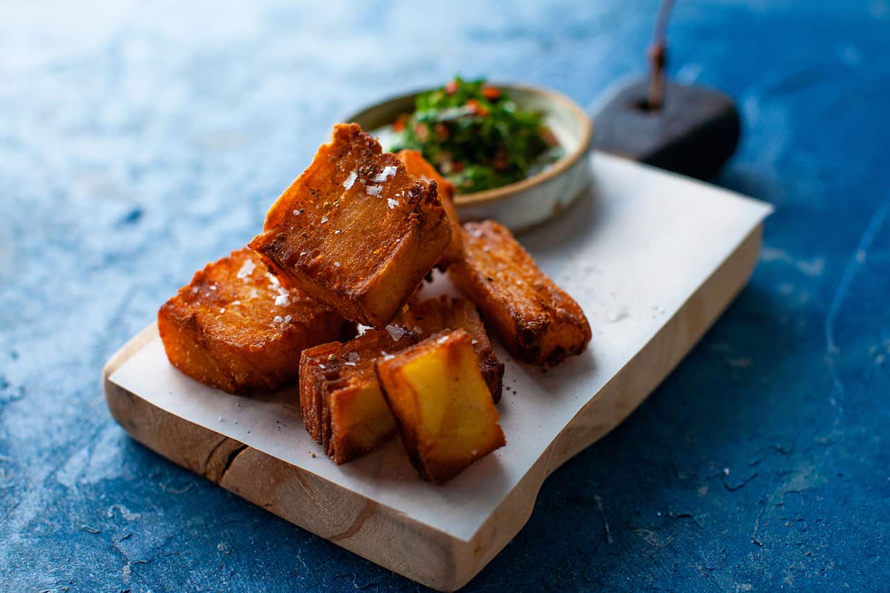 A pile of crunchy, dark golden brown potato chunks sit on a board, along with a bowl of green sauce. 