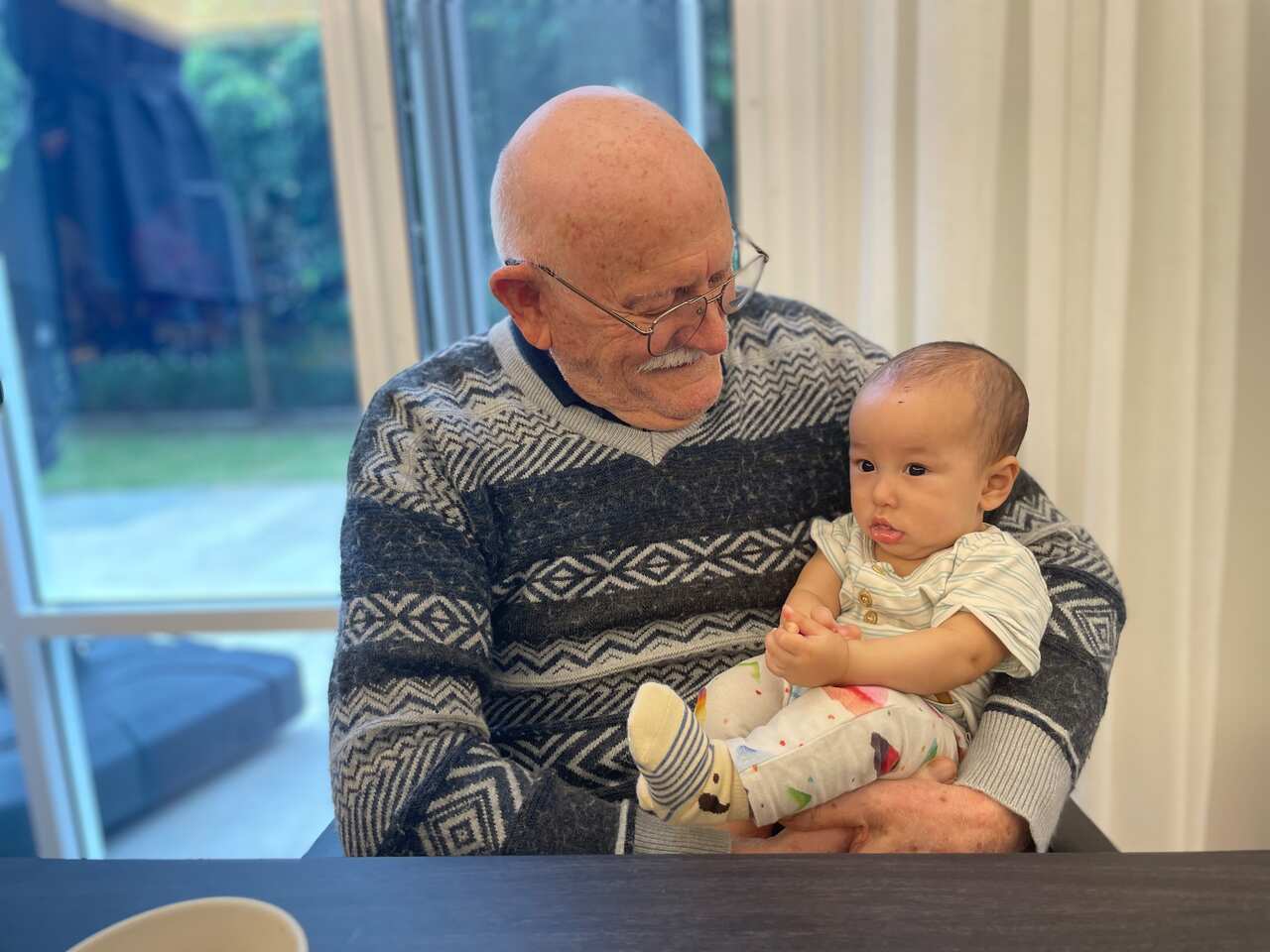 An older man carries a baby in his lap on a dining table. 