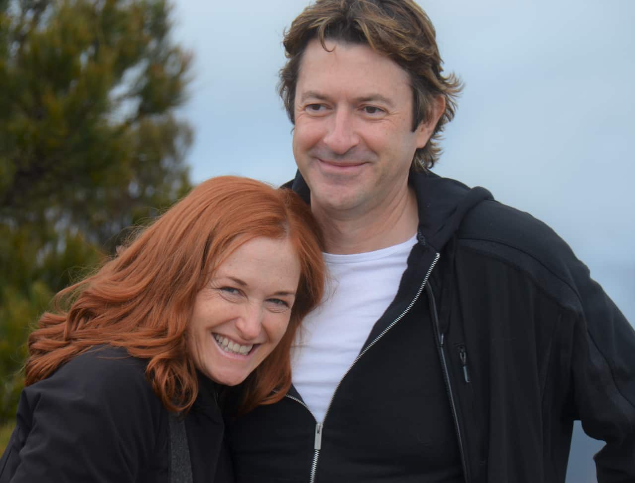 A man in a black zipper is standing outside next to a smiling woman with auburn hair. 
