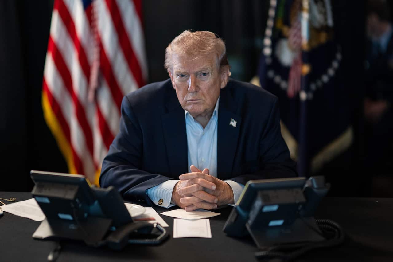 Donald Trump stares into the camera as he sits at his desk flanked by two phones and the US flag behind him.