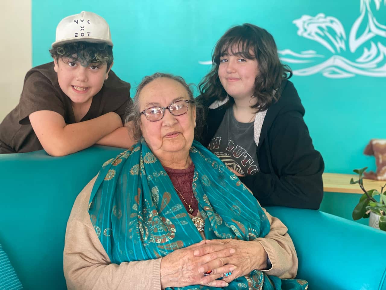 Colleen Prideaux sits on a chair, wearing glasses and a blue shawl, with her grandchildren either side of her