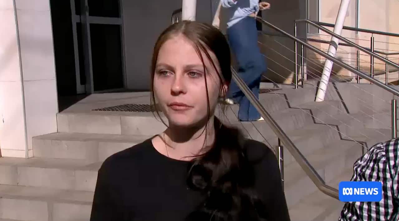 A young woman with dark hair in a black top stands outside a courthouse with eyes red from crying. 