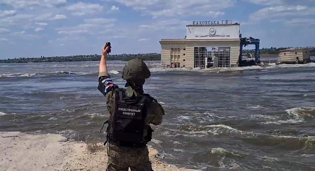 Man clad in army uniform holds object in left hand to the sky as flood waters envelop a building in the distance.  