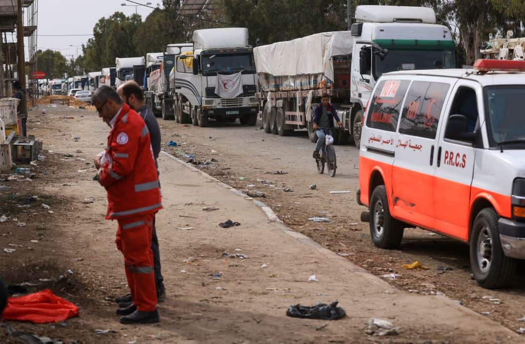 A line of trucks on a dusty road. Two men stand praying by the side of the road.